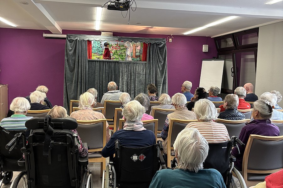Gruppe älterer Menschen sitzt in einem Raum mit lila Wänden und schaut auf eine Bühne mit Puppentheater
