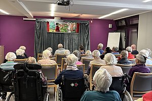Gruppe älterer Menschen sitzt in einem Raum mit lila Wänden und schaut auf eine Bühne mit Puppentheater