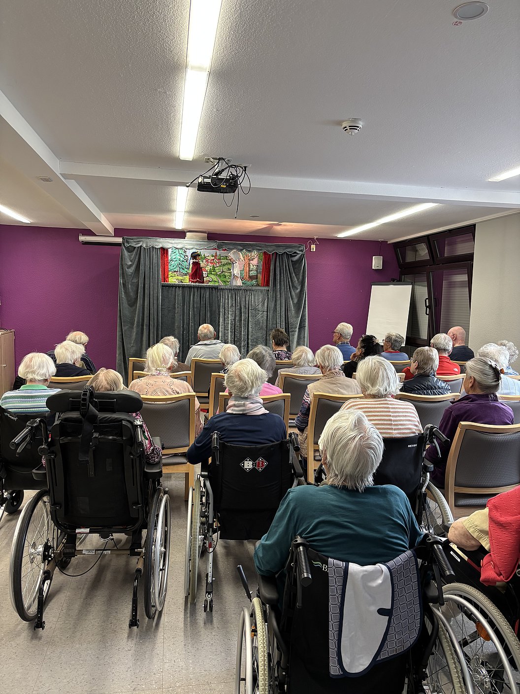 Gruppe älterer Menschen sitzt in einem Raum mit lila Wänden und schaut auf eine Bühne mit Puppentheater