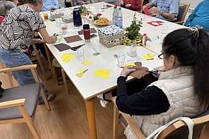 Mehrere ältere Menschen sitzen an einem Tisch und arbeiten mit Papier und Stiften