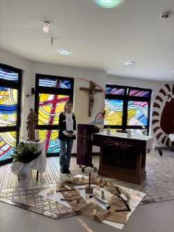 Person steht in einem Raum mit bunten Glasfenstern, einem Altar und einem großen Kreuz an der Wand